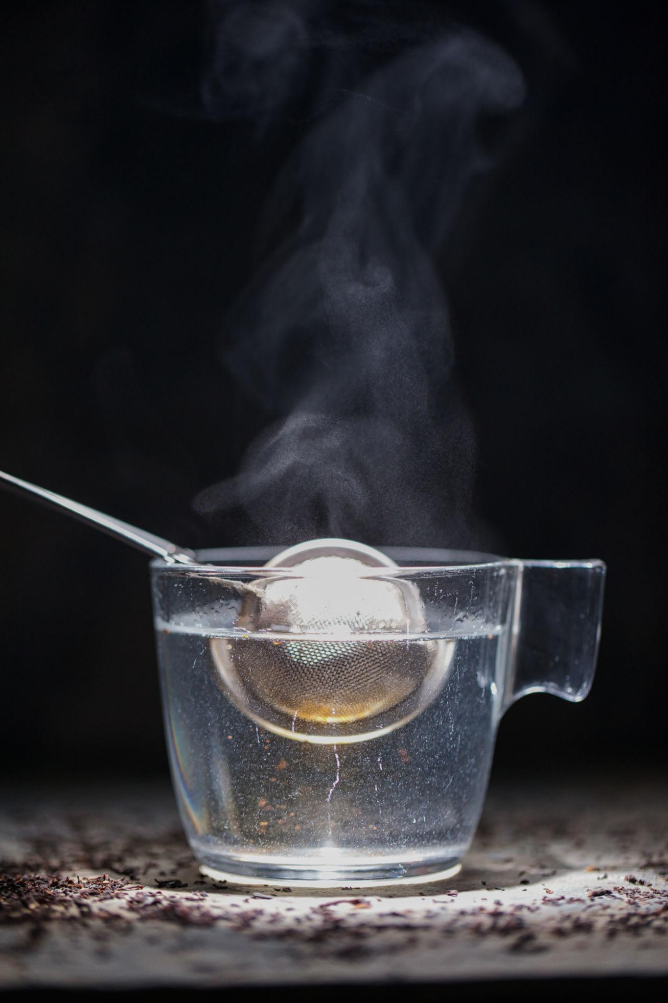 Verre d’eau chaude et boule à thé en inox, vapeur sur fond sombre.